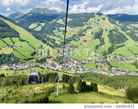 Aerial view on Saalbach village and mountains in Saalbach-Hinterglemm skiing region in Austria on a beautiful summer day from cableway on the village with holiday ressorts, the church and other Aerial view on Saalbach village and mountains in Saalbach-Hinterglemm skiing region in Austria on a beautiful summer day from cableway on the village with holiday ressorts, the church and other 68893455