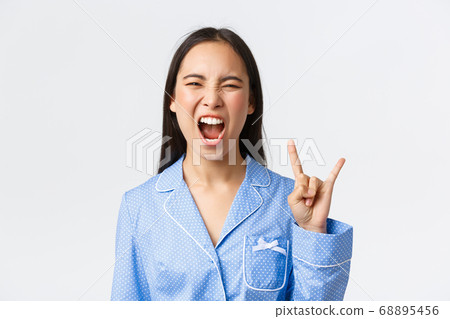 Close-up of rebellious and carefree attractive asian girl in blue pajama having slumber party, showing rock-n-roll gesture as going wild, shouting yeah and enjoying music, white background 68895456