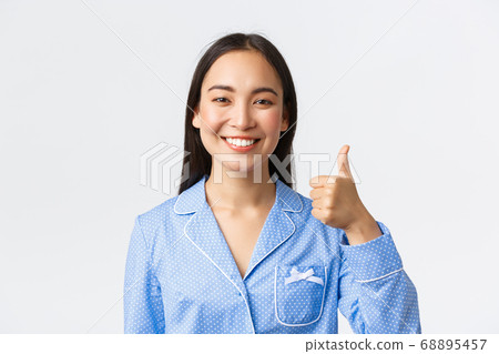 Close-up of satisfied pretty smiling asian woman in blue pajama showing thumbs-up in approval, recommend and guarantee product quality, standing pleased over white background 68895457