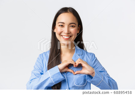 Adorable happy asian girl in blue pajama love staying at home, wearing cozy jammies, showing heart gesture and smiling delighted, standing white background upbeat 68895458