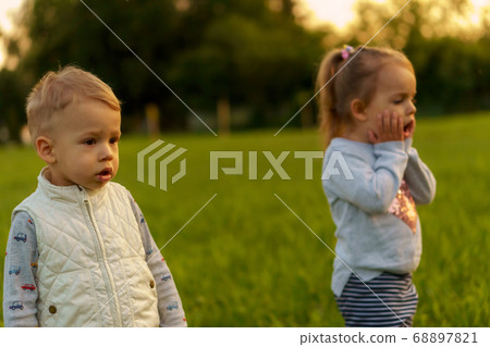 infancy, childhood, development, autumn mood, medicine and health concept - two small children boy and girl of same age, demonstrate emotions of fear and surprise on green meadow in park at sunset. infancy, childhood, development, autumn mood, medicine and health concept - two small children boy and girl of same age, demonstrate emotions of fear and surprise on green meadow in park at sunset. 68897821
