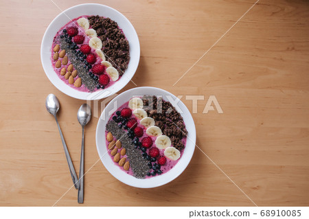 Smoothie bowl with fresh raspberry, blueberry, chia seeds, almonds, banana, and chocolate flakes. Morning breakfast on wooden table. Top view of two plates. vegan food Smoothie bowl with fresh raspberry, blueberry, chia seeds, almonds, banana, and chocolate flakes. Morning breakfast on wooden table. Top view of two plates. vegan food 68910085