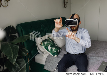 young man with a beard in virtual reality glasses sits on the edge of the bed 68917992