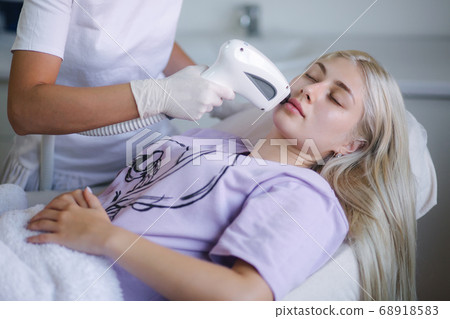 Cosmetology. Beautiful woman receiving laser hair removal procedure at beauty salon. Beautician hands doing beauty treatment for female face at spa salon. Facial Treatment Cosmetology. Beautiful woman receiving laser hair removal procedure at beauty salon. Beautician hands doing beauty treatment for female face at spa salon. Facial Treatment 68918583