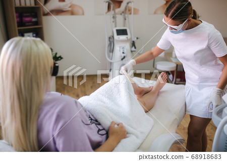 Young woman beautician in mask applies contact gel on the leg of a woman for laser hair removal in medical clinic Young woman beautician in mask applies contact gel on the leg of a woman for laser hair removal in medical clinic 68918683