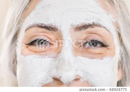 Close up beauty cropped portrait of charming gray haired senior lady, with white facial mask on face, looking at camera while standing on isolated light background. Skin care for elderly women Close up beauty cropped portrait of charming gray haired senior lady, with white facial mask on face, looking at camera while standing on isolated light background. Skin care for elderly women 68928775