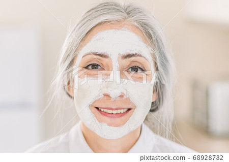 Skin care and anti age beauty treatments, copy space. Indoor shot of beautiful retired grau haired woman with clay moisturizing mask on her face, standing on light kitchen background at home 68928782