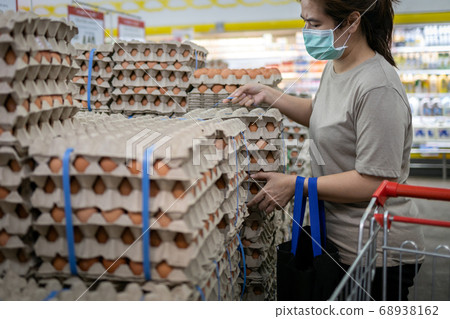 Asian woman wearing face mask,choosing fresh chicken eggs while shopping food,people panic buying and  hoarding during the Covid-19, Coronavirus epidemic.girl preparing for pathogen virus pandemic 68938162