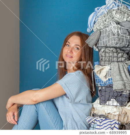 Young red-haired woman sitting on the wooden floor with stack of wardrobe and linen in metal laundry basket. 68939995