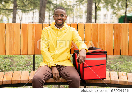 Young cheerful male courier in yellow jacket keeping hand on big red backpack 68954966