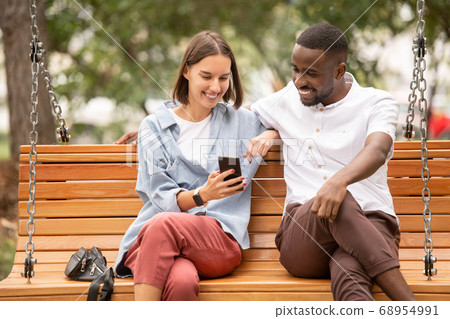 Young smiling woman showing her African boyfriend curious video in smartphone 68954991