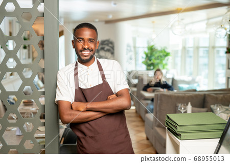Cheerful young African waiter in workwear leaning against door in restaurant 68955013