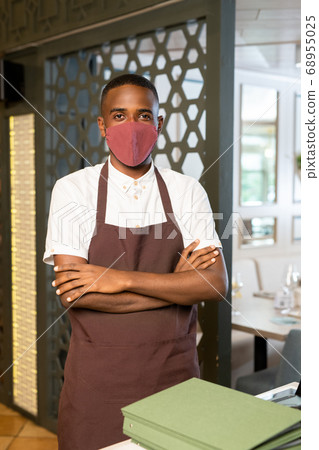 Contemporary waiter in protective mask and brown apron crossing arms on chest Contemporary waiter in protective mask and brown apron crossing arms on chest 68955025