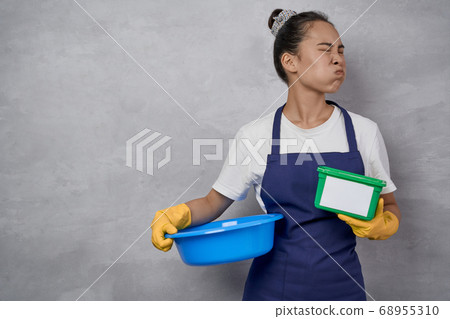 Portrait of unhappy housewife or maid woman in uniform holding basin and green plastic box with washing capsules, making unsatisfied face while standing against grey wall Portrait of unhappy housewife or maid woman in uniform holding basin and green plastic box with washing capsules, making unsatisfied face while standing against grey wall 68955310