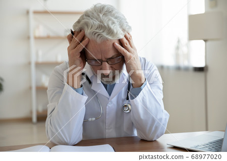 Exhausted unhappy mature doctor wearing glasses sitting at desk 68977020