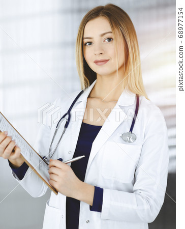 Young optimistic woman-doctor is holding a clipboard in her hands, while standing in a sunny clinic. Portrait of friendly female physician with a stethoscope. Perfect medical service in a hospital Young optimistic woman-doctor is holding a clipboard in her hands, while standing in a sunny clinic. Portrait of friendly female physician with a stethoscope. Perfect medical service in a hospital 68977154