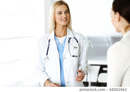 Woman-doctor at work in sunny hospital is happy to consult female patient. Blonde physician checks medical history record and exam results while using clipboard Woman-doctor at work in sunny hospital is happy to consult female patient. Blonde physician checks medical history record and exam results while using clipboard 68982662