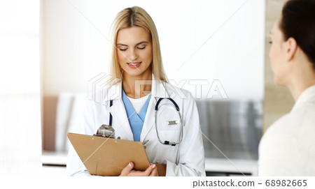 Woman-doctor at work in sunny hospital is happy to consult female patient. Blonde physician checks medical history record and exam results while using clipboard Woman-doctor at work in sunny hospital is happy to consult female patient. Blonde physician checks medical history record and exam results while using clipboard 68982665