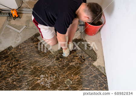 The man sets a tile of dark brown on the glue The man sets a tile of dark brown on the glue 68982834