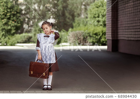 a little girl in a retro school uniform and a white apron walks down the street with a briefcase after school. a little girl in a retro school uniform and a white apron walks down the street with a briefcase after school. 68984430