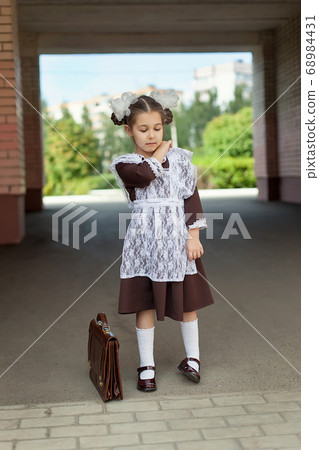 a little girl on the street in the uniform of the USSR with a briefcase stands near the school. Day of knowledge. a little girl on the street in the uniform of the USSR with a briefcase stands near the school. Day of knowledge. 68984431
