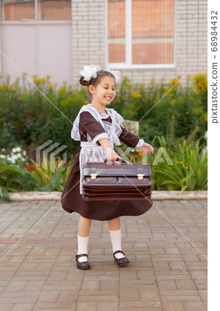 a little girl on the street in the uniform of the USSR with a briefcase stands near the school. Day of knowledge. a little girl on the street in the uniform of the USSR with a briefcase stands near the school. Day of knowledge. 68984432
