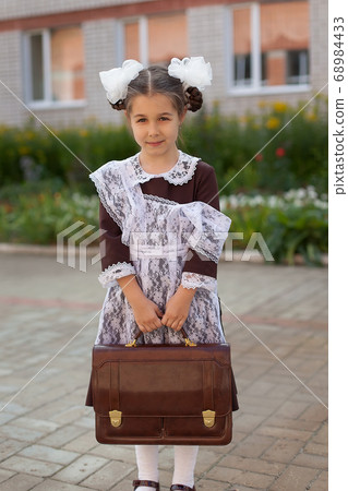 a little girl on the street in the uniform of the USSR with a briefcase stands near the school. Day of knowledge. 68984433
