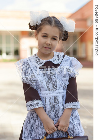 a little girl in a retro school uniform and a white apron walks down the street with a briefcase after school. a little girl in a retro school uniform and a white apron walks down the street with a briefcase after school. 68984453