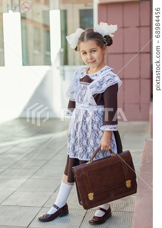 a little girl in a retro school uniform and a white apron walks down the street with a briefcase after school. a little girl in a retro school uniform and a white apron walks down the street with a briefcase after school. 68984456