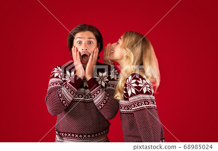 Shocked millennial guy in Christmas outfit and girl whispering something into his ear on red background Shocked millennial guy in Christmas outfit and girl whispering something into his ear on red background 68985024