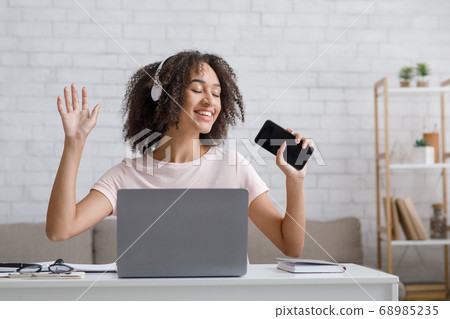 Take a break from work at home. Happy african american woman with headphones singing at smartphone Take a break from work at home. Happy african american woman with headphones singing at smartphone 68985235