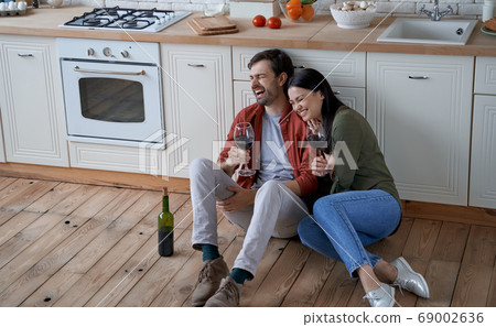 Romantic evening at home. Young happy romantic couple, wife and husband sitting on the floor in the modern kitchen, drinking wine and hugging 69002636