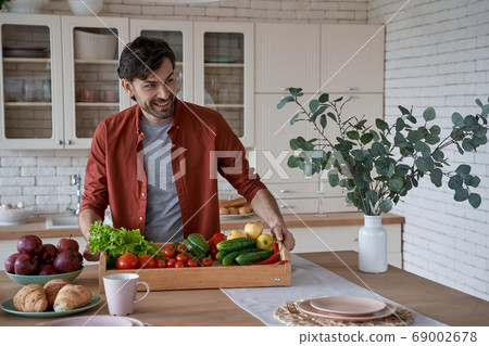 Vegetarianism concept. Happy young man in casual clothes holding basket with various fresh vegetables and smiling, cooking healthy food at home 69002678