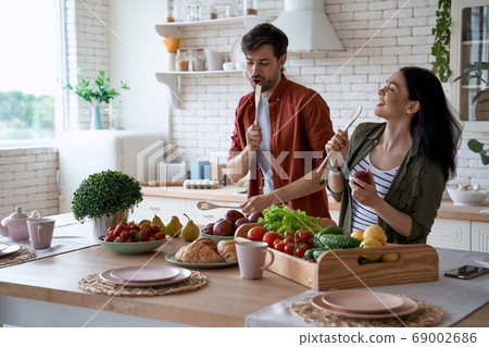 Young happy family couple dancing, singing and having fun while preparing healthy food in the modern kitchen at home. Fresh fruits and vegetables on the wooden table Young happy family couple dancing, singing and having fun while preparing healthy food in the modern kitchen at home. Fresh fruits and vegetables on the wooden table 69002686