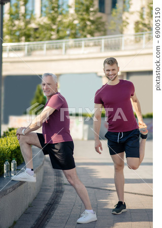 Mature man stretching knee, bearded male stretching quad next to him, both smiling Mature man stretching knee, bearded male stretching quad next to him, both smiling 69005150