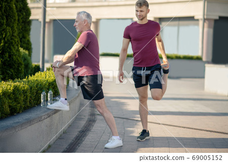 Grey-haired man stretching knee, young male stretching quad next to him Grey-haired man stretching knee, young male stretching quad next to him 69005152