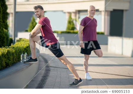 Young male stretching knee, grey-haired man stretching quad behind him Young male stretching knee, grey-haired man stretching quad behind him 69005153