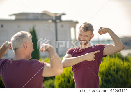 Bearded male showing biceps to grey-haired man, smiling Bearded male showing biceps to grey-haired man, smiling 69005161