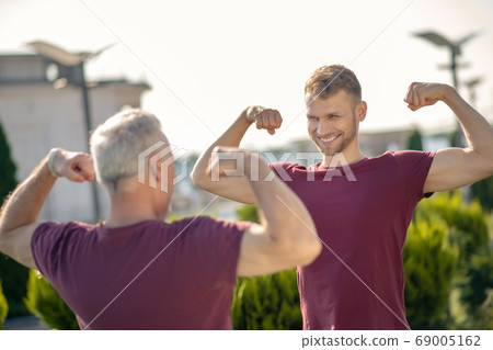 Bearded male and grey-haired standing opposite, showing biceps, smiling Bearded male and grey-haired standing opposite, showing biceps, smiling 69005162