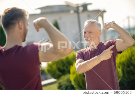 Young male and grey-haired man standing, showing each other biceps, smiling Young male and grey-haired man standing, showing each other biceps, smiling 69005163