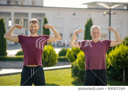 Bearded young male and mature grey-haired man standing, showing biceps Bearded young male and mature grey-haired man standing, showing biceps 69005164