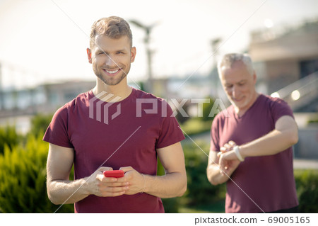 Bearded male holding smartphone, smiling, grey-haired man verifying smartwatch behind him Bearded male holding smartphone, smiling, grey-haired man verifying smartwatch behind him 69005165