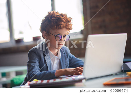 Red-haired boy sitting at the laptop and looking busy Red-haired boy sitting at the laptop and looking busy 69006474