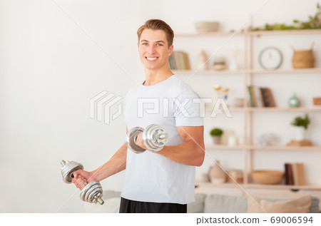 Strength training at home during quarantine. Smiling handsome guy lifts dumbbells 69006594