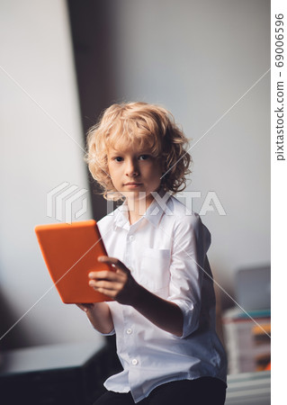 Cute blonde boy holding his tablet and looking serious Cute blonde boy holding his tablet and looking serious 69006596