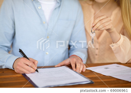 Cropped view of young couple with house key signing real estate purchase agreement at table, close up Cropped view of young couple with house key signing real estate purchase agreement at table, close up 69006961