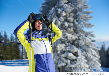 Happy woman posing on skis before skiing. Sunny day at ski resort. Clear blue sky, snow-covered fir trees on background. 69013312