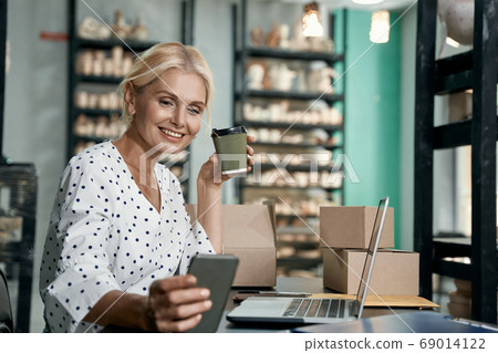 Successful business owner. Beautiful and happy business woman drinking coffee, using smartphone and smiling while working in her art studio or craft pottery shop Successful business owner. Beautiful and happy business woman drinking coffee, using smartphone and smiling while working in her art studio or craft pottery shop 69014122