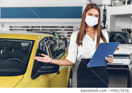 Woman car seller standing near new car wearing protective face mask Woman car seller standing near new car wearing protective face mask 69015036