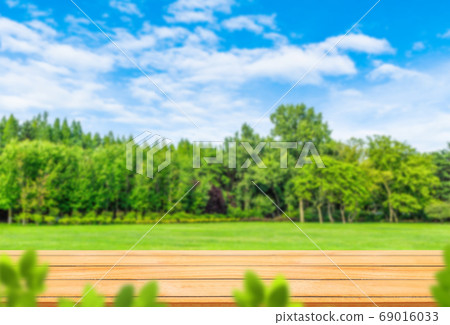 Wooden table top with Blurry image of green grass meadow field and green trees. Wooden table top with Blurry image of green grass meadow field and green trees. 69016033